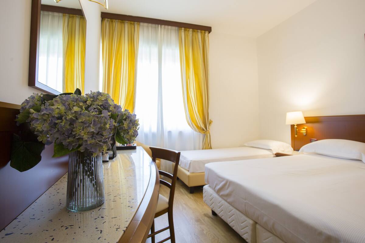 A hotel room with a bed, a desk with a vase of flowers, and a window with curtains.