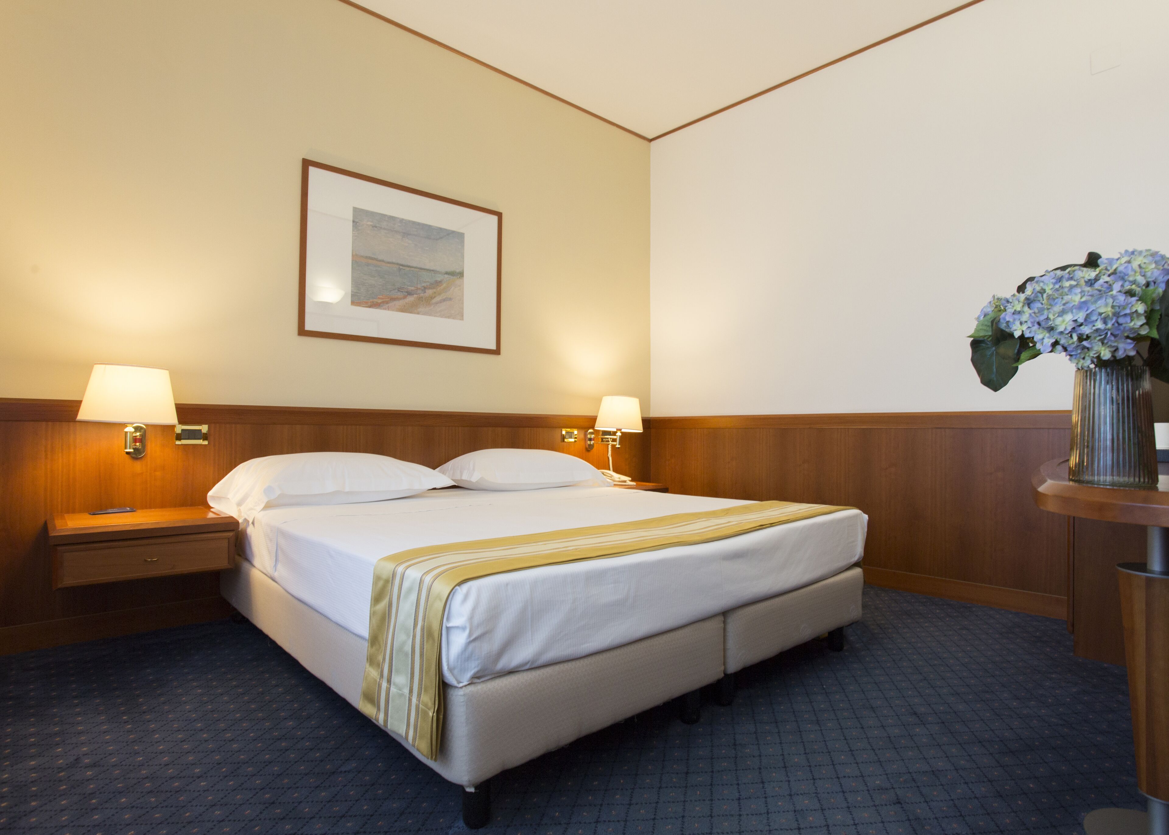 A hotel room with a bed, two bedside lamps, a nightstand, a framed picture on the wall, and a vase with flowers on a side table.