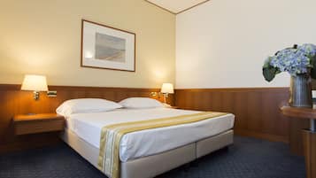 Habitación de hotel con cama, dos lámparas de noche, una mesita de noche, un cuadro enmarcado en la pared y un florero con flores sobre una mesita auxiliar.