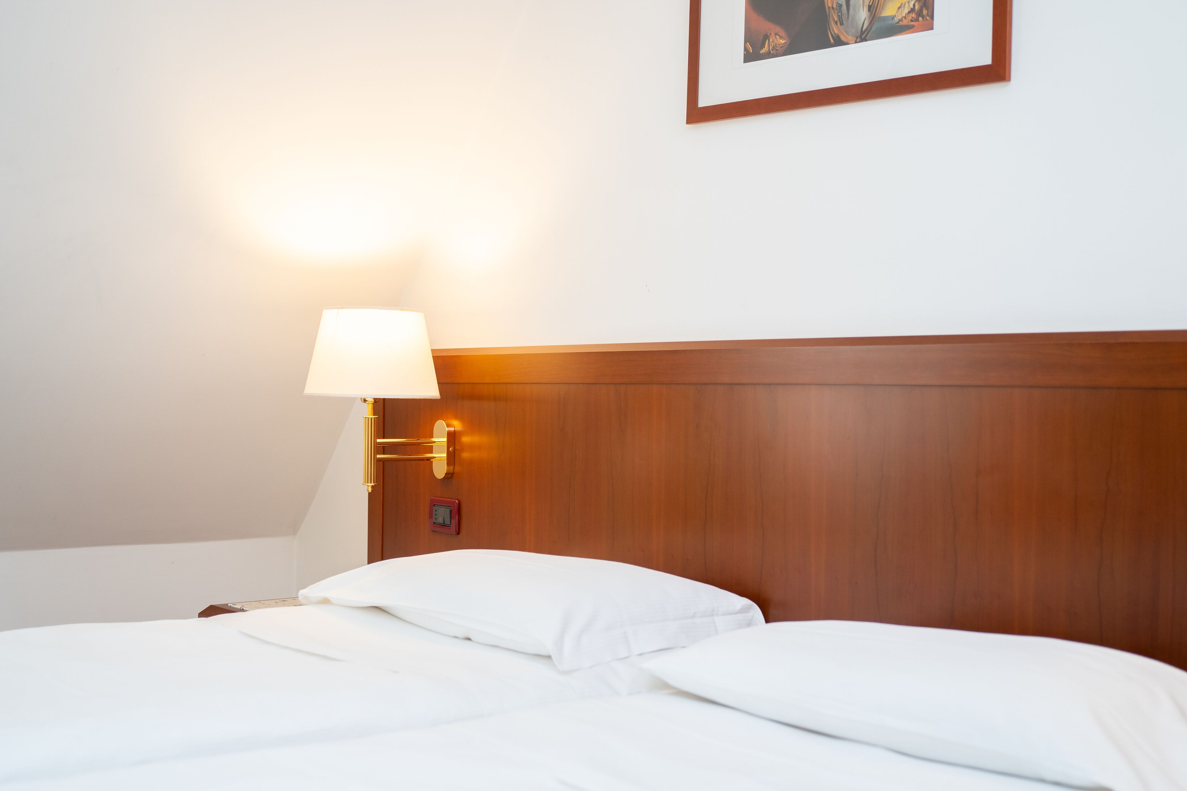 Una cama bien hecha con sábanas blancas, cabecera de madera y una lámpara fijada a la pared.