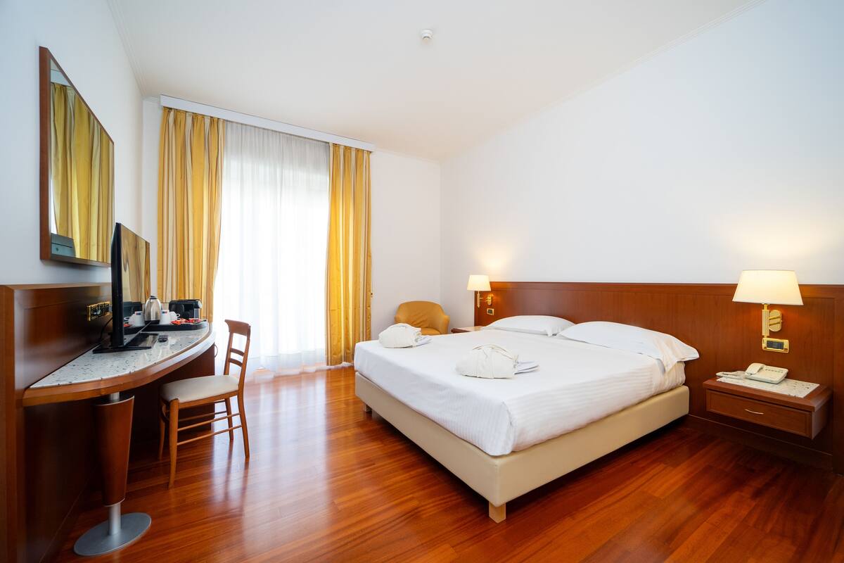 A hotel room with a bed, a desk, a chair, a nightstand, and a lamp.