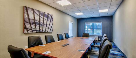 A conference room with a long wooden table, black chairs, a large abstract painting on the wall, and a window providing natural light.