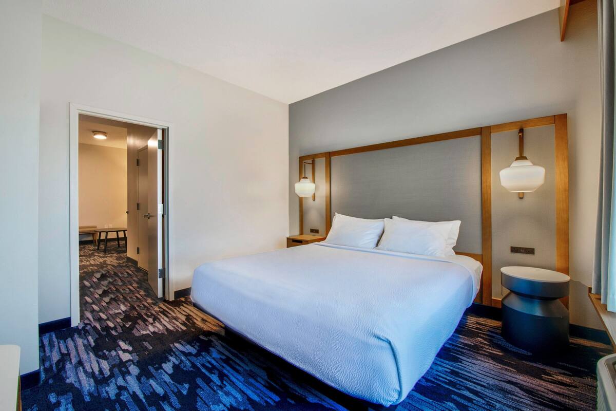 A hotel room with a large bed, a bedside table, a nightstand, a lamp, and a carpeted floor.