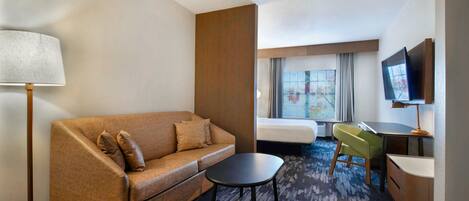 A hotel room with a sofa, a small table, a desk with a chair, a TV, and a window with curtains.