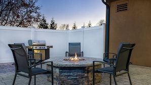 Een terras met een vuurkorf van steen, een barbecue en twee stoelen.