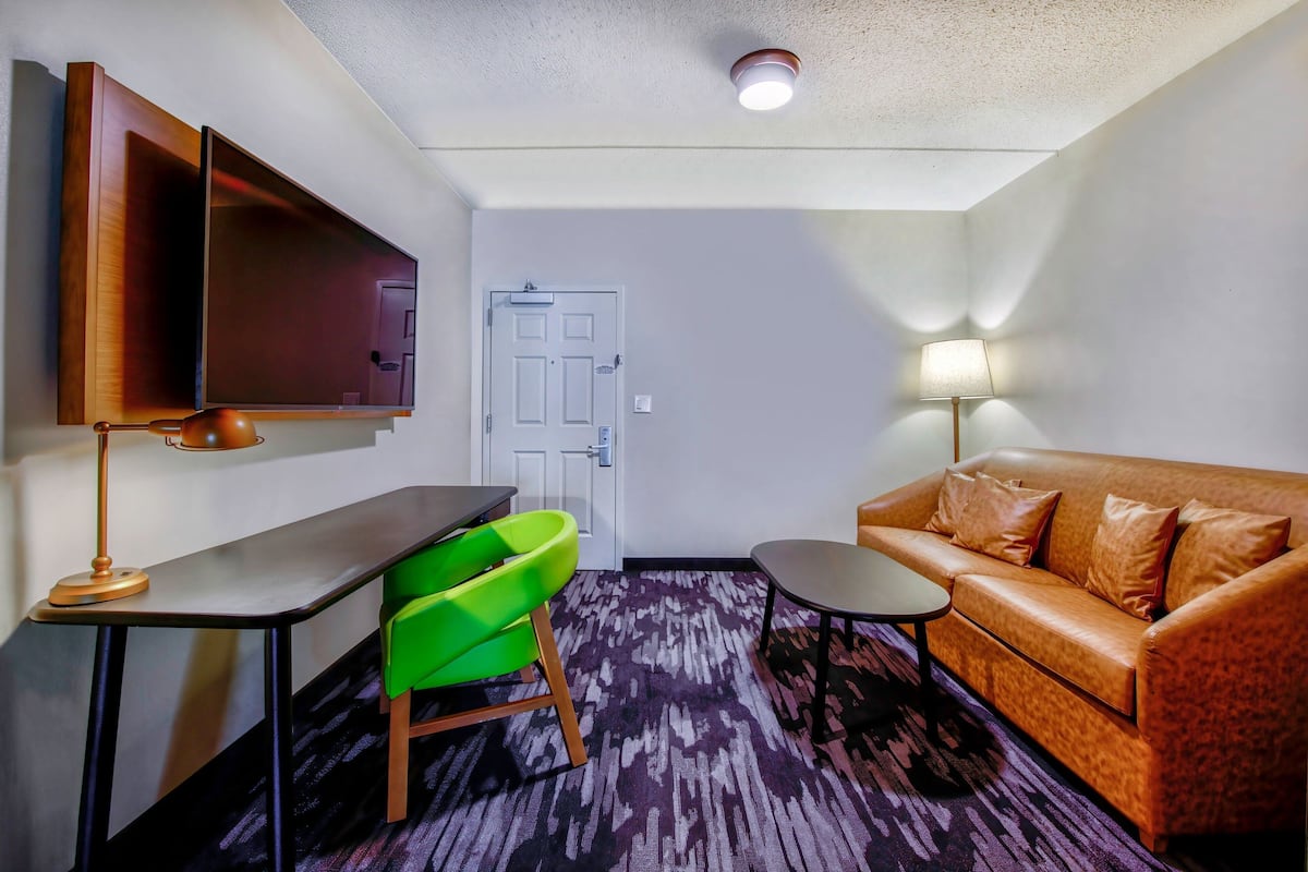 A compact room with a sofa, a small table, a desk with a chair, a television, and a door.