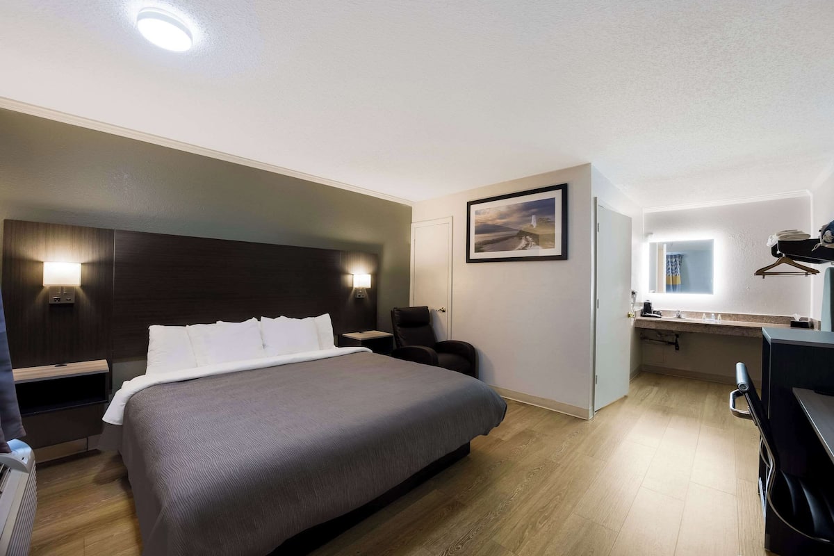 A hotel room with a large bed, a desk, a chair, and a small kitchenette.