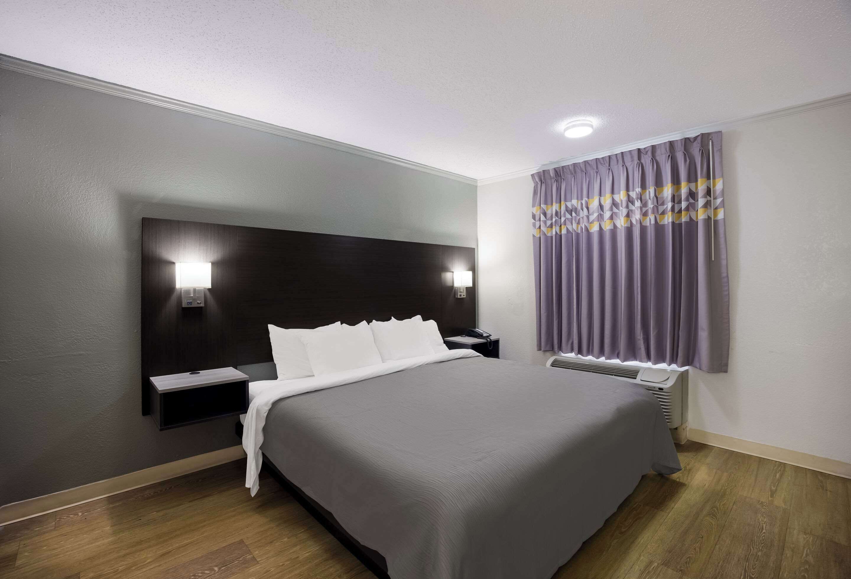 A modern hotel room with a large bed, bedside tables, a built-in heater, and a window with curtains.