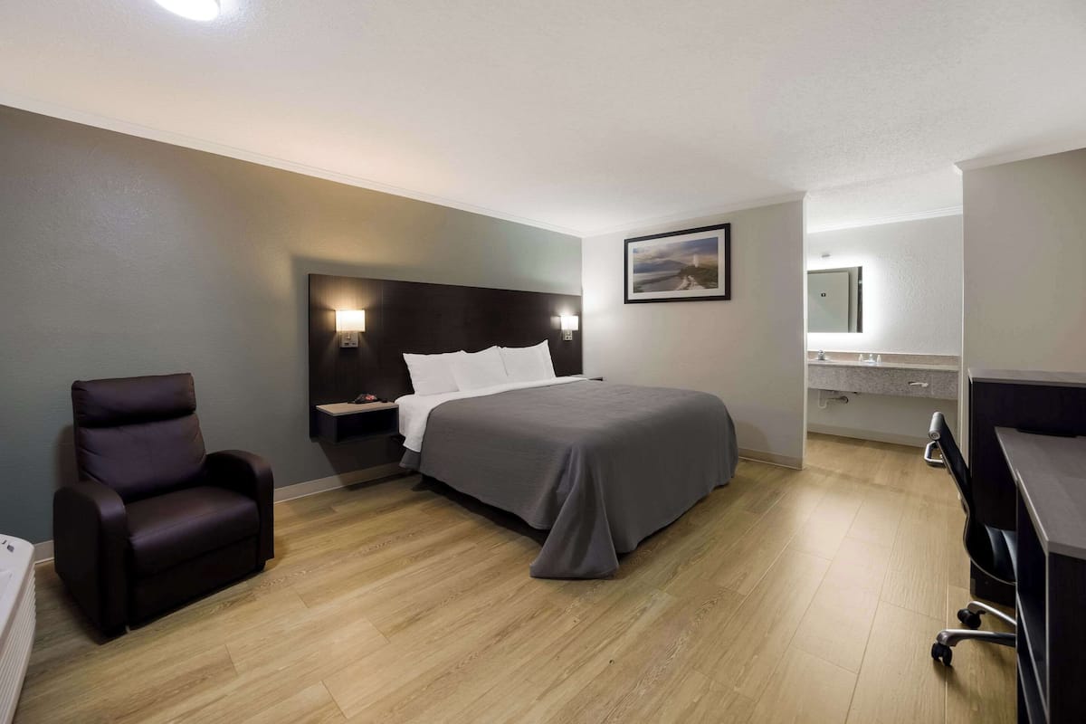 A modern hotel room with a bed, a chair, a desk, and a bathroom mirror.