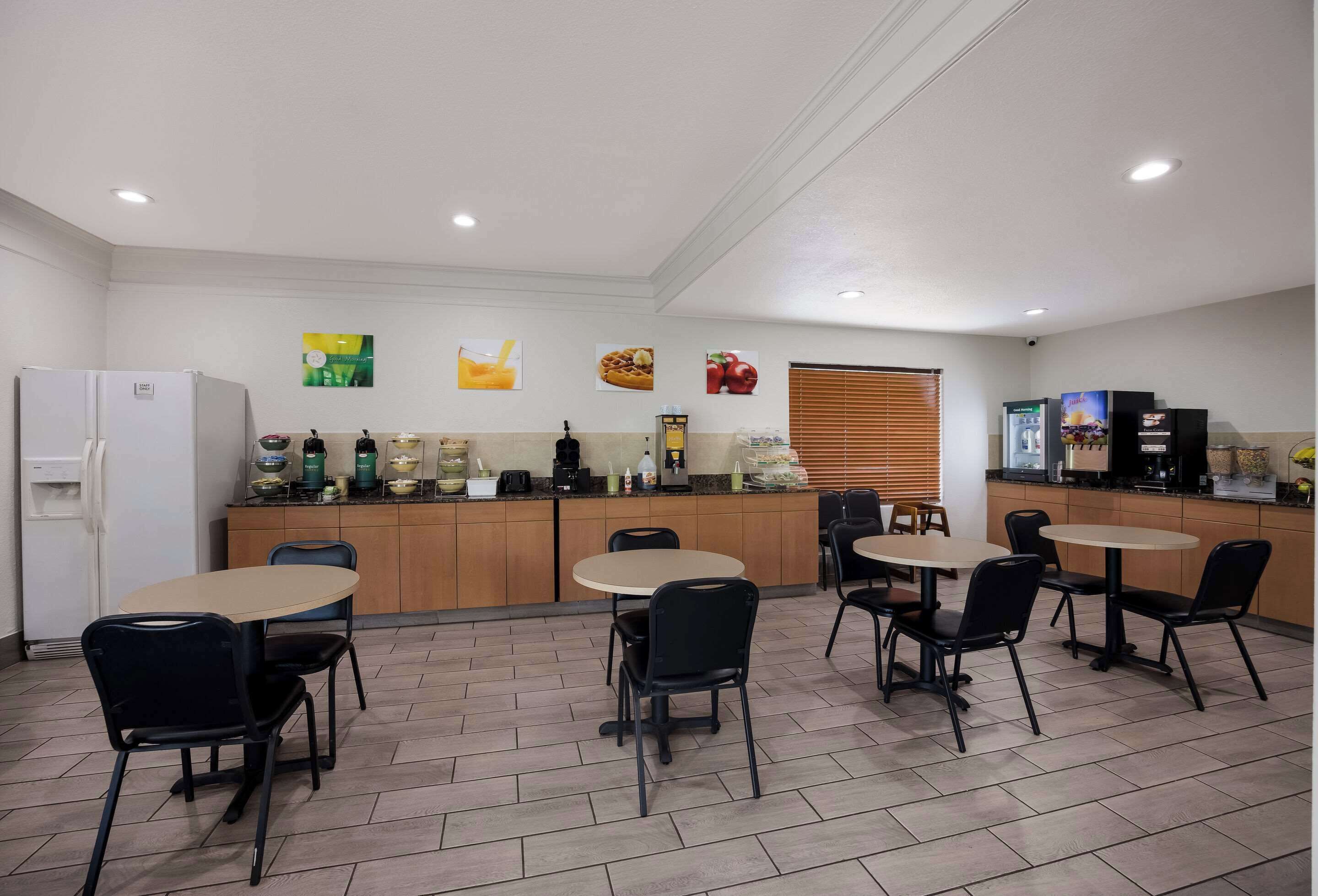 Uma sala de jantar limpa e moderna, com bancada de cozinha, várias mesas e cadeiras, e uma geladeira.