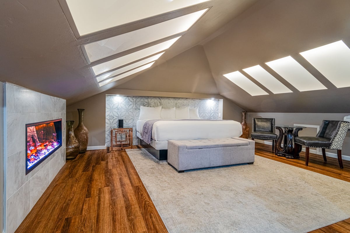 A modern bedroom with a skylight, a bed, a bench, and a fireplace.