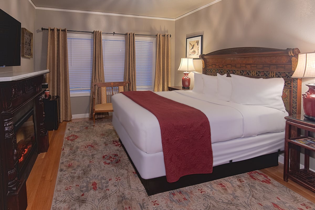 A hotel room with a large bed, a wooden headboard, a nightstand with a lamp, a chair, a fireplace, and a window with curtains.
