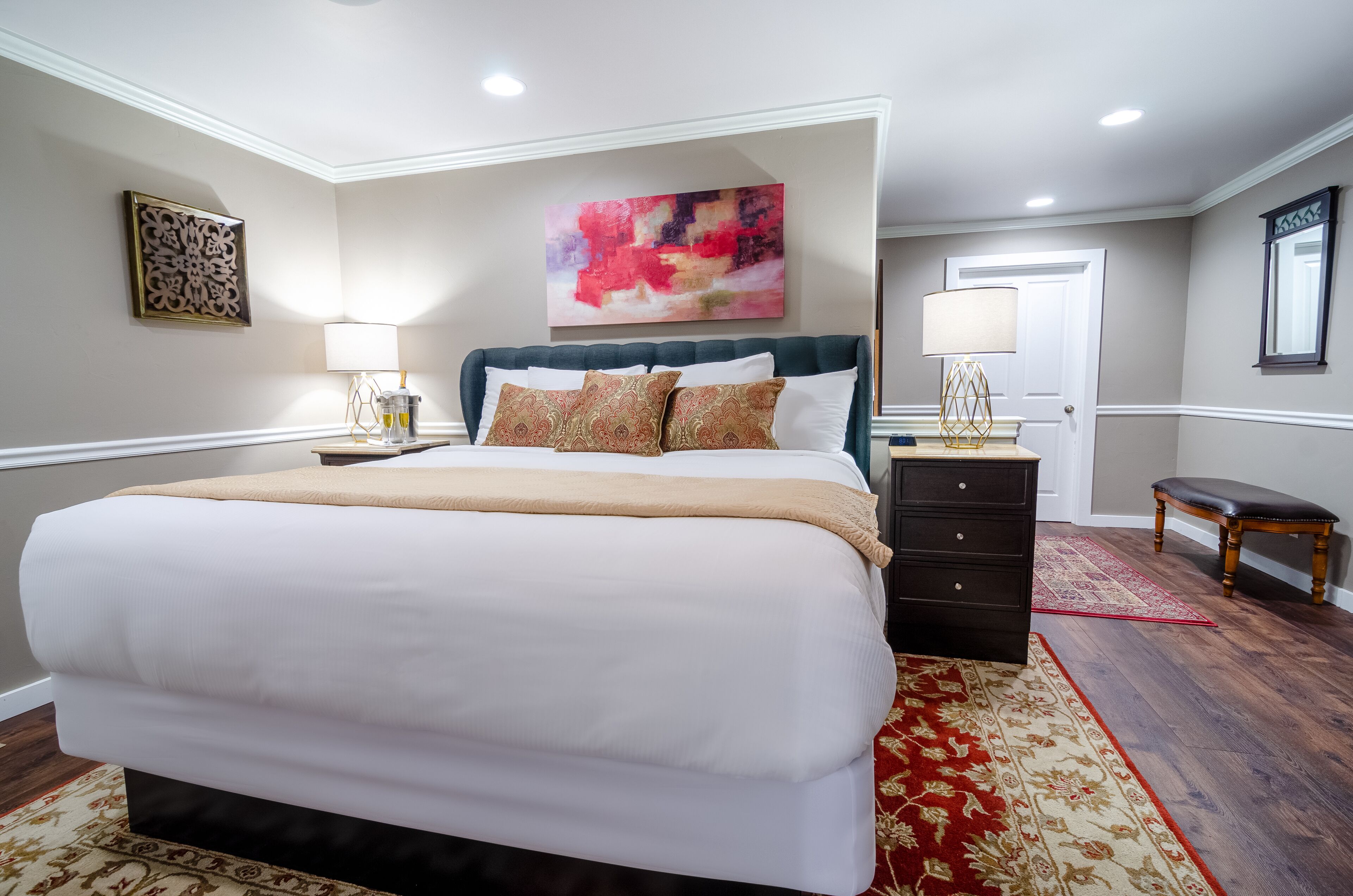 A bedroom with a large bed, a dresser, a bench, and a painting on the wall.
