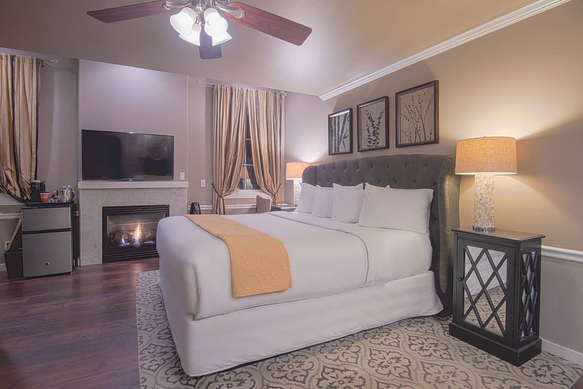 A hotel room with a large bed, a television, a fireplace, and a side table with a lamp.