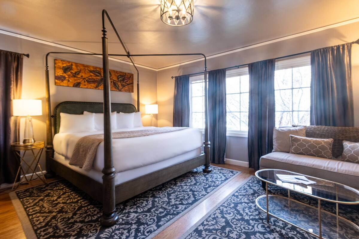 A four-poster bed with a wooden frame, a patterned rug, a sofa, and a glass coffee table in a well-lit room with large windows and curtains.