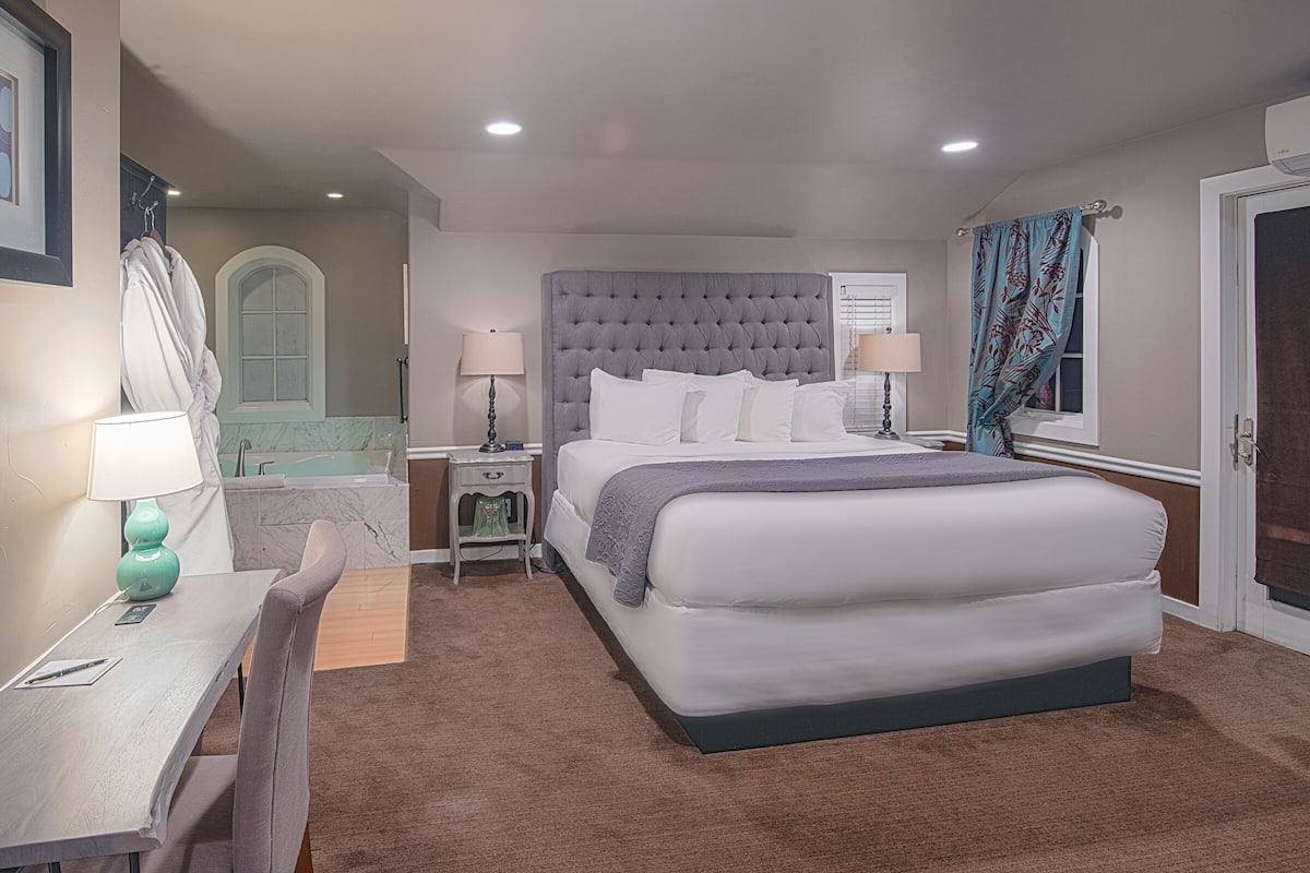 A hotel room with a large bed, two bedside tables with lamps, a desk with a chair, a bathroom with a mirror and sink, and a door with a curtain.