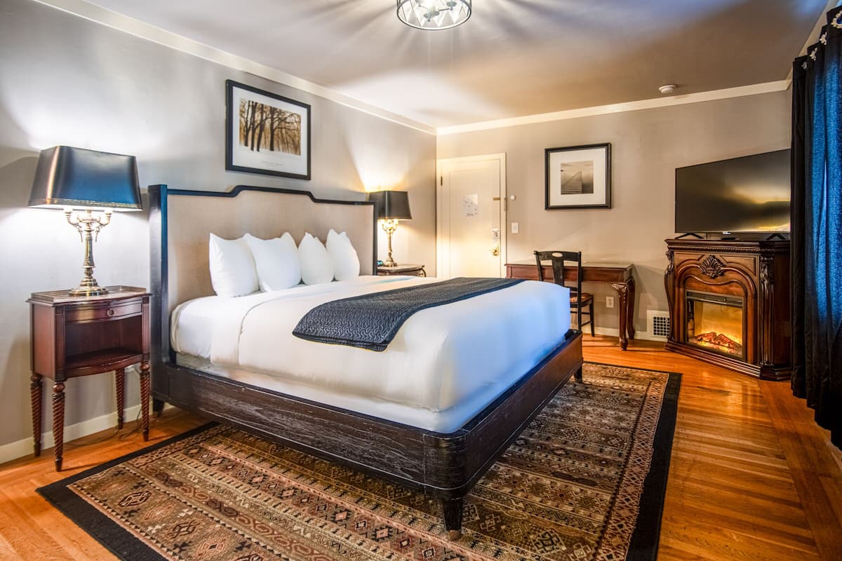 A hotel room with a large bed, two bedside tables, a TV, a fireplace, and a desk.