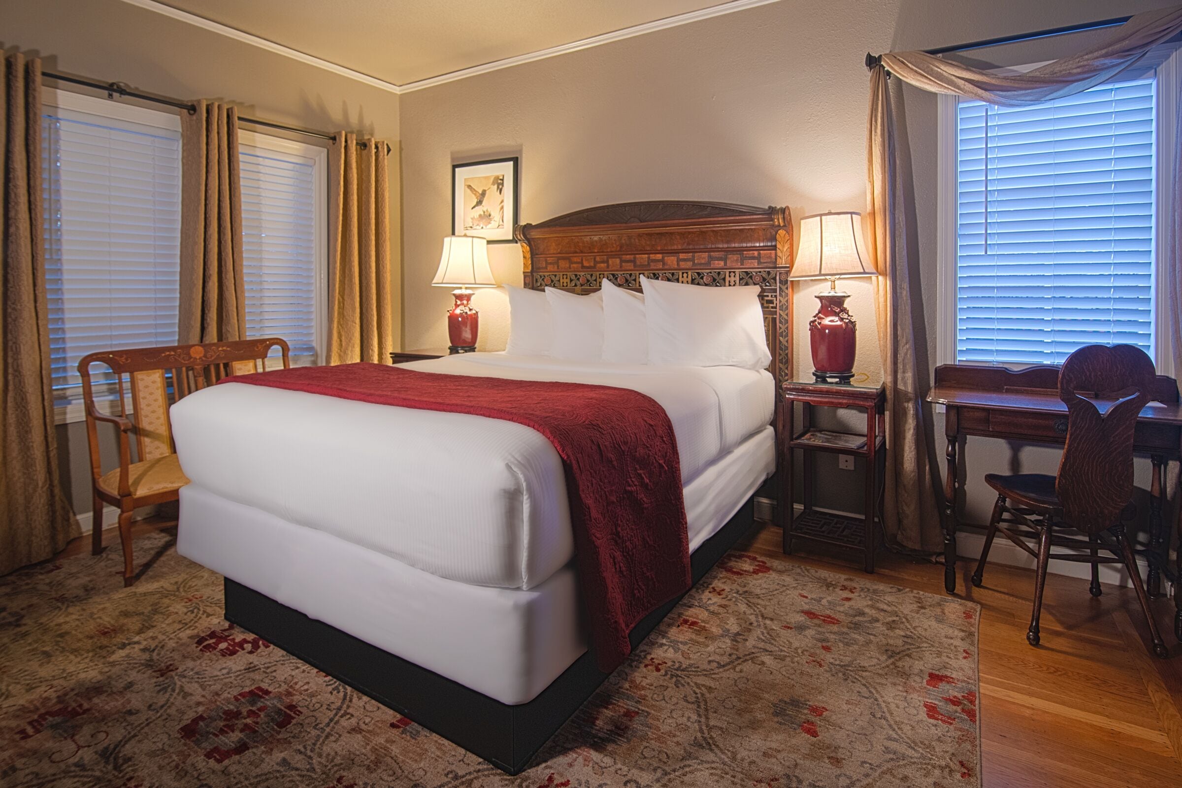 A hotel room with a large bed, a desk with a chair, a nightstand with lamps, and a window with curtains.