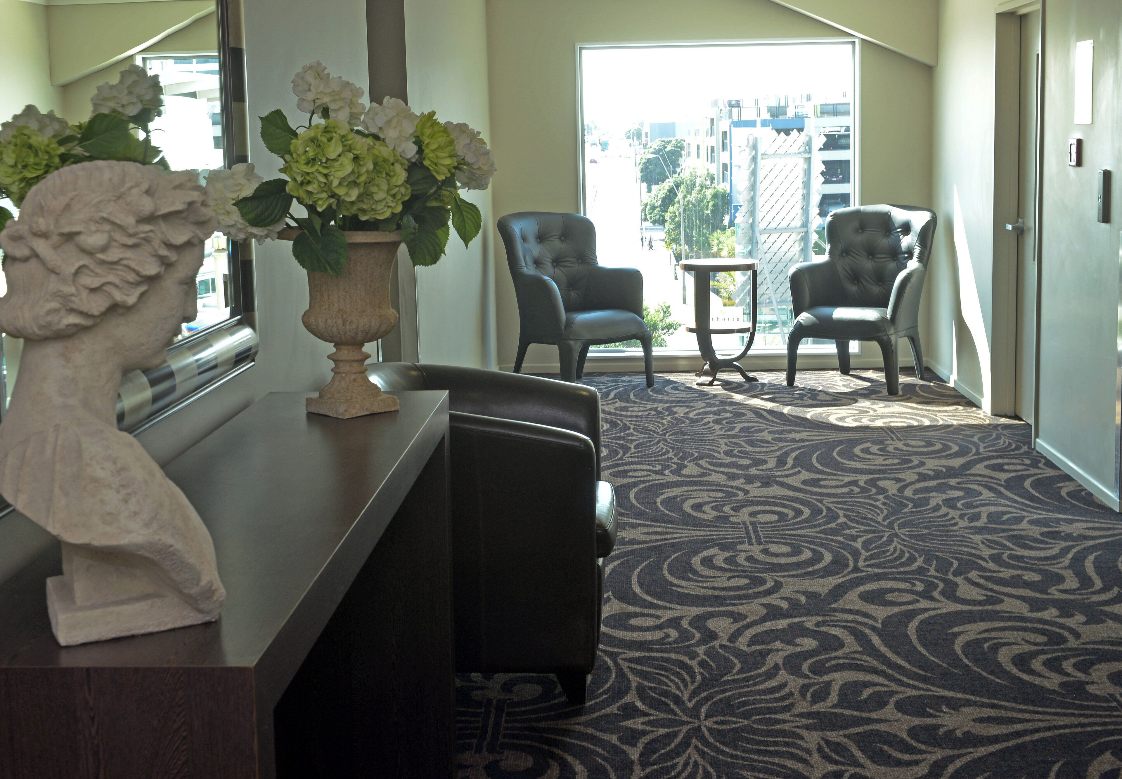 A hotel lobby with a reception desk, two armchairs, a small table, and a patterned carpet.
