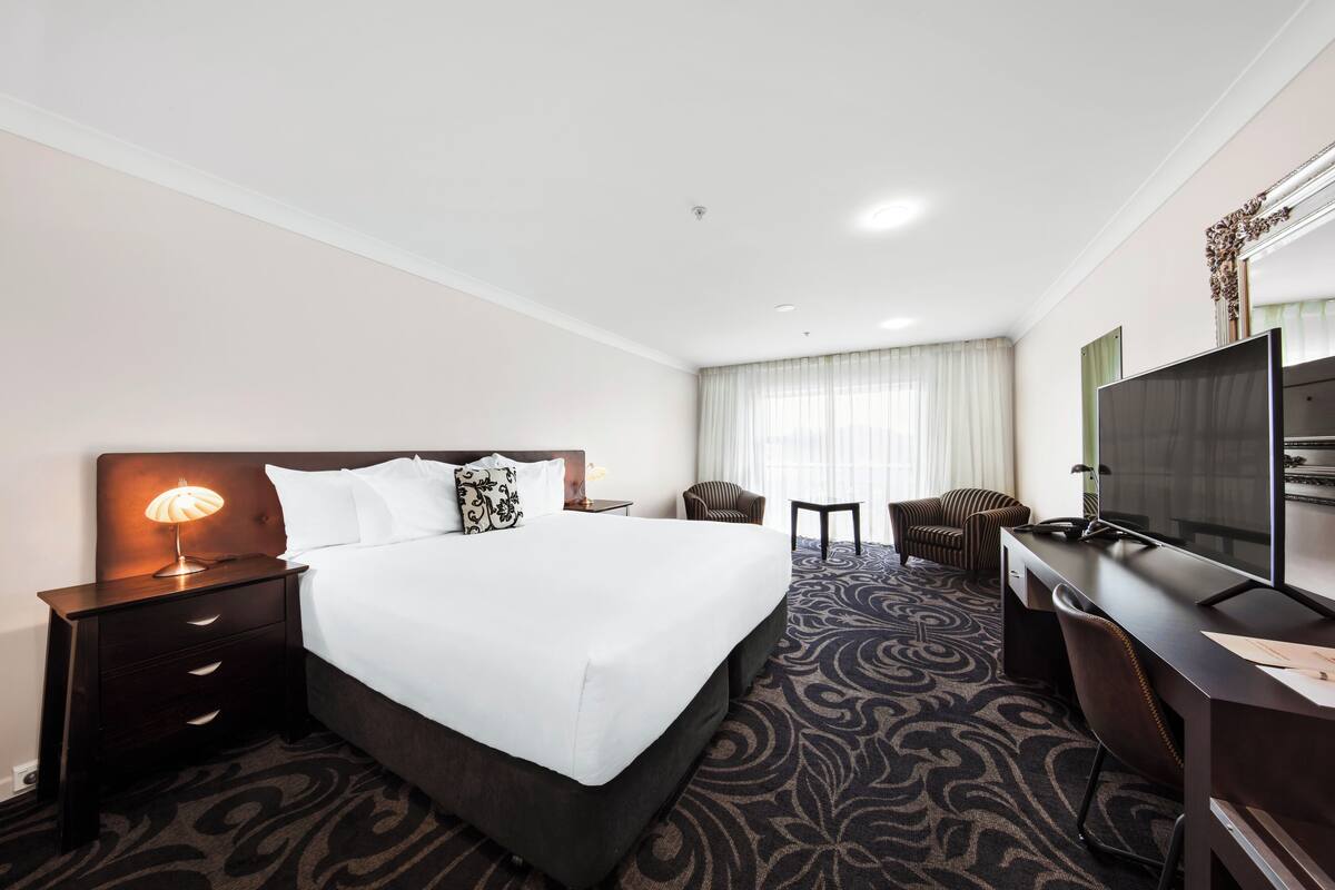 Una habitación de hotel con una cama grande, un escritorio con computadora, un televisor, una silla y una lámpara.