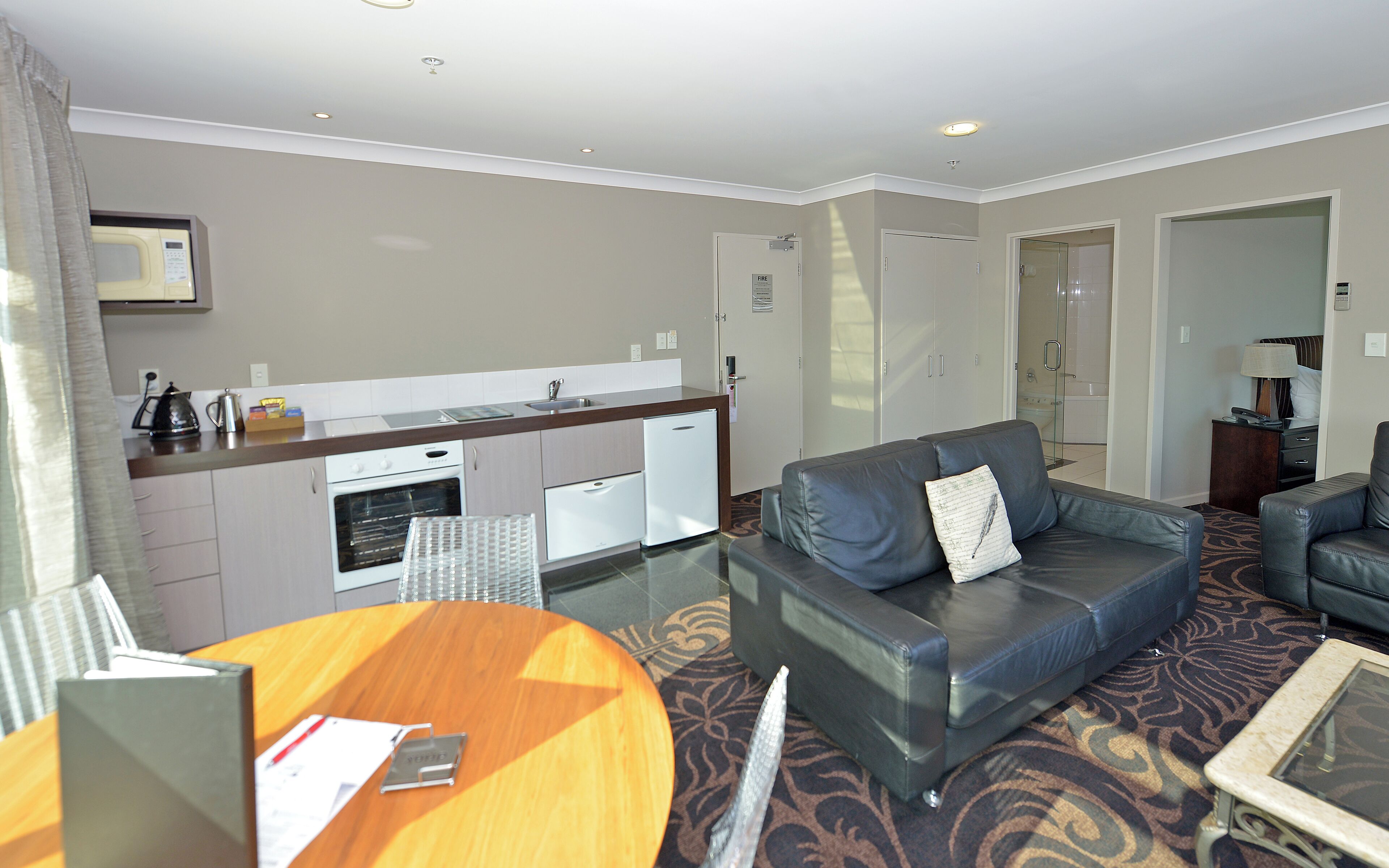 A hotel room with a kitchenette, a leather sofa, a round wooden table, and a glass shower area.