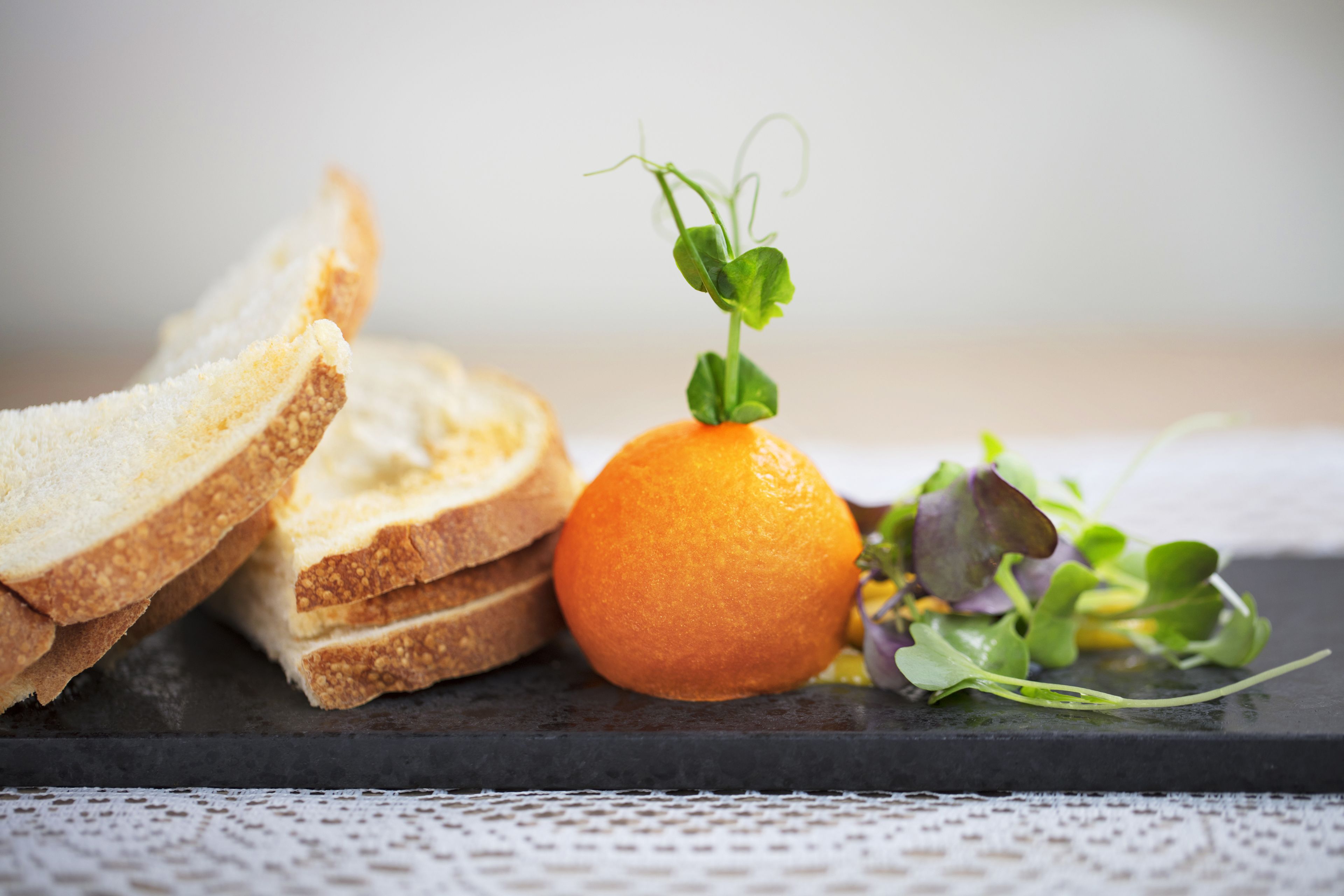 A black slate serving board with slices of bread, an orange, and a small bunch of greens.