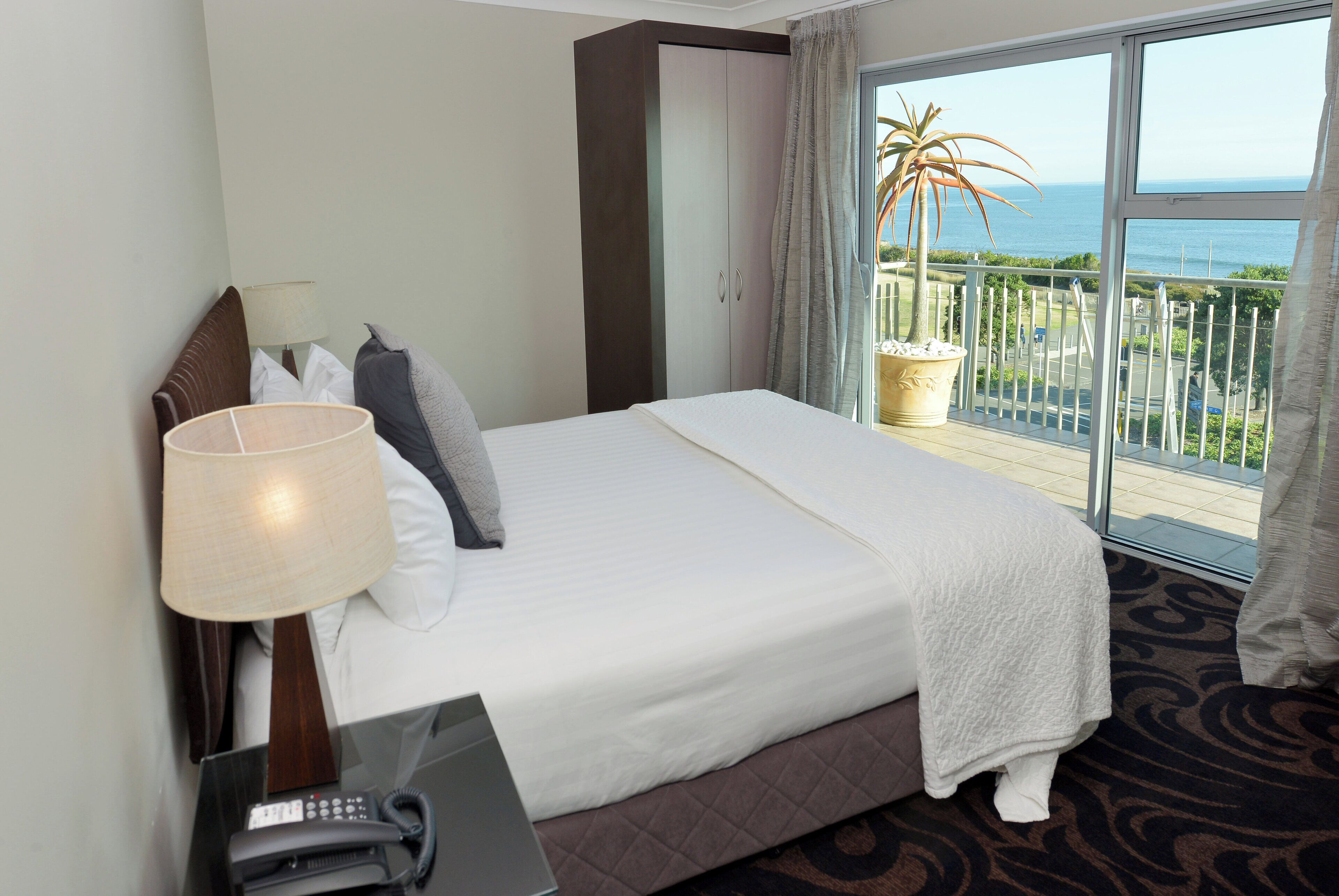 A hotel room with a bed, a desk with a telephone, and a view of the ocean through a balcony.