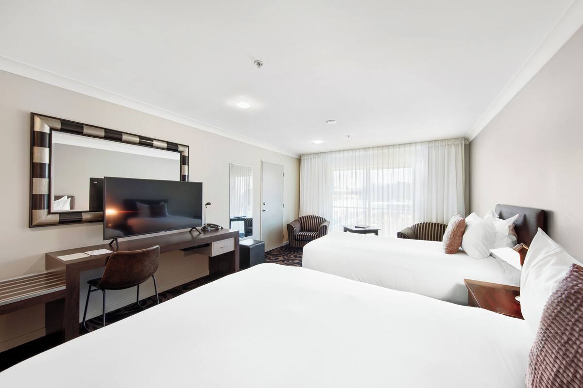 Habitación de hotel con dos camas, un escritorio, una silla, un televisor y un espejo grande.
