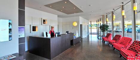 Et moderne hotelværelse med reception, receptionist, skrivebord og rødt mønstret siddemøbel.