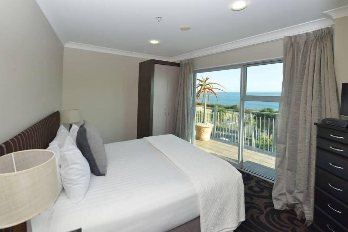 Habitación de hotel con cama, tocador, lámpara y balcón con vista al mar.