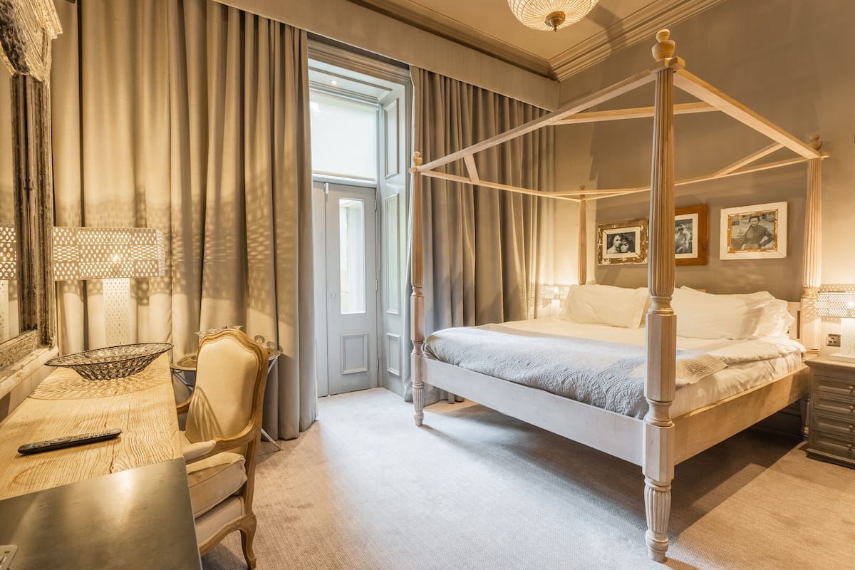Ein Bett mit Baldachin, ein Schreibtisch aus Holz mit Lampe, ein Sessel und ein Fenster mit Vorhängen.