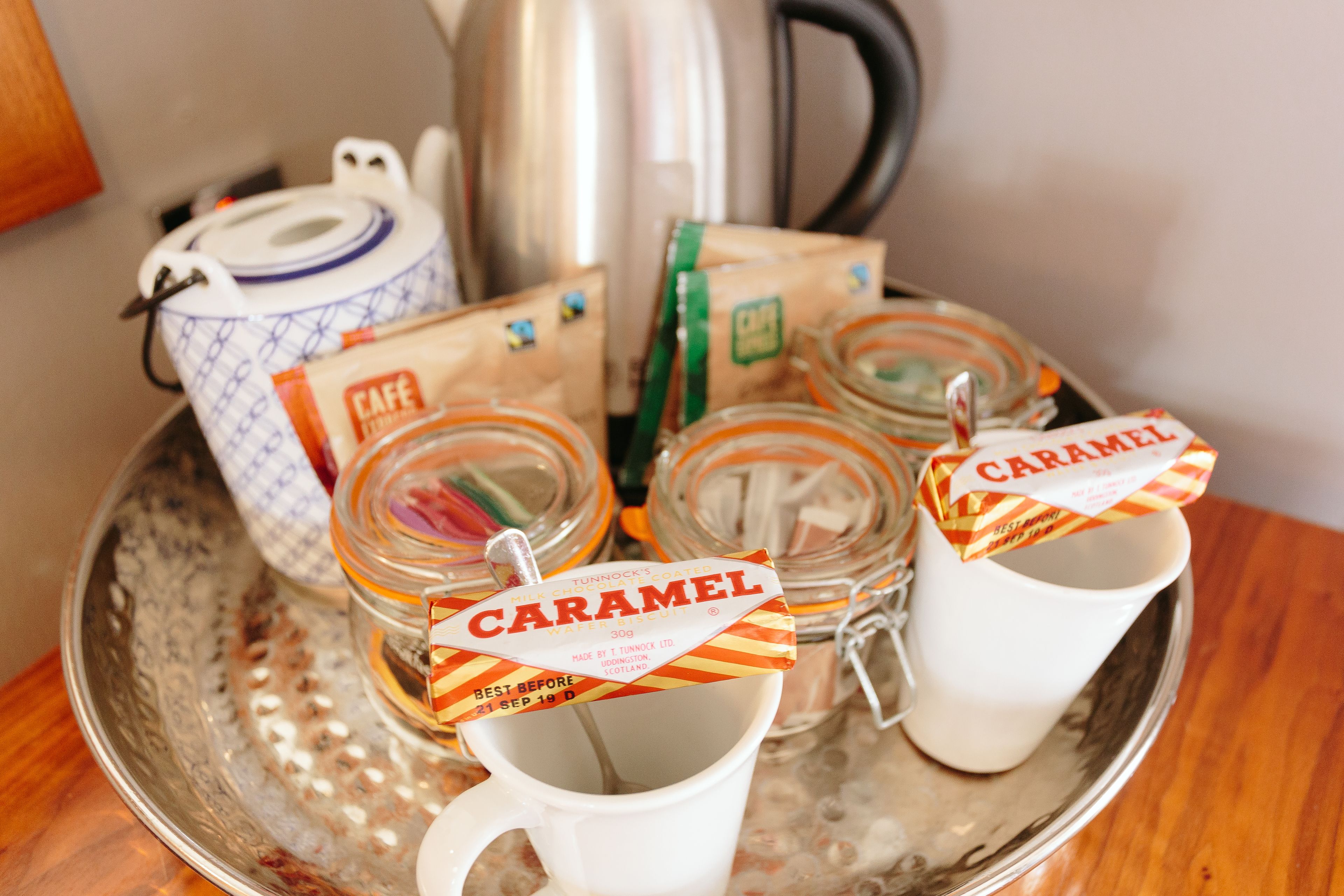A tray with jars labeled "CARAMEL" and a white mug, a blue and white teapot, and a silver kettle on a metallic tray.