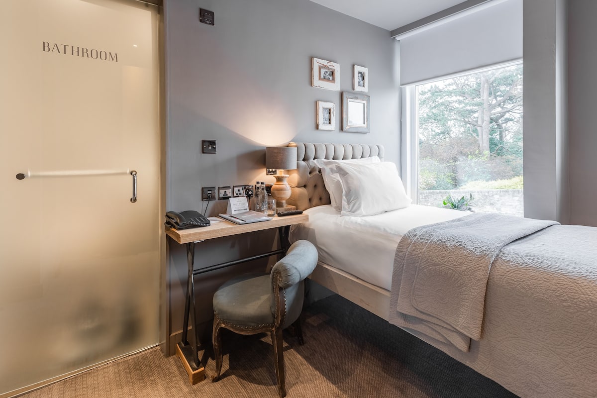 Ein Schlafzimmer mit Bett, Schreibtisch mit Lampe, Stuhl und Blick auf Bäume durch ein Fenster.