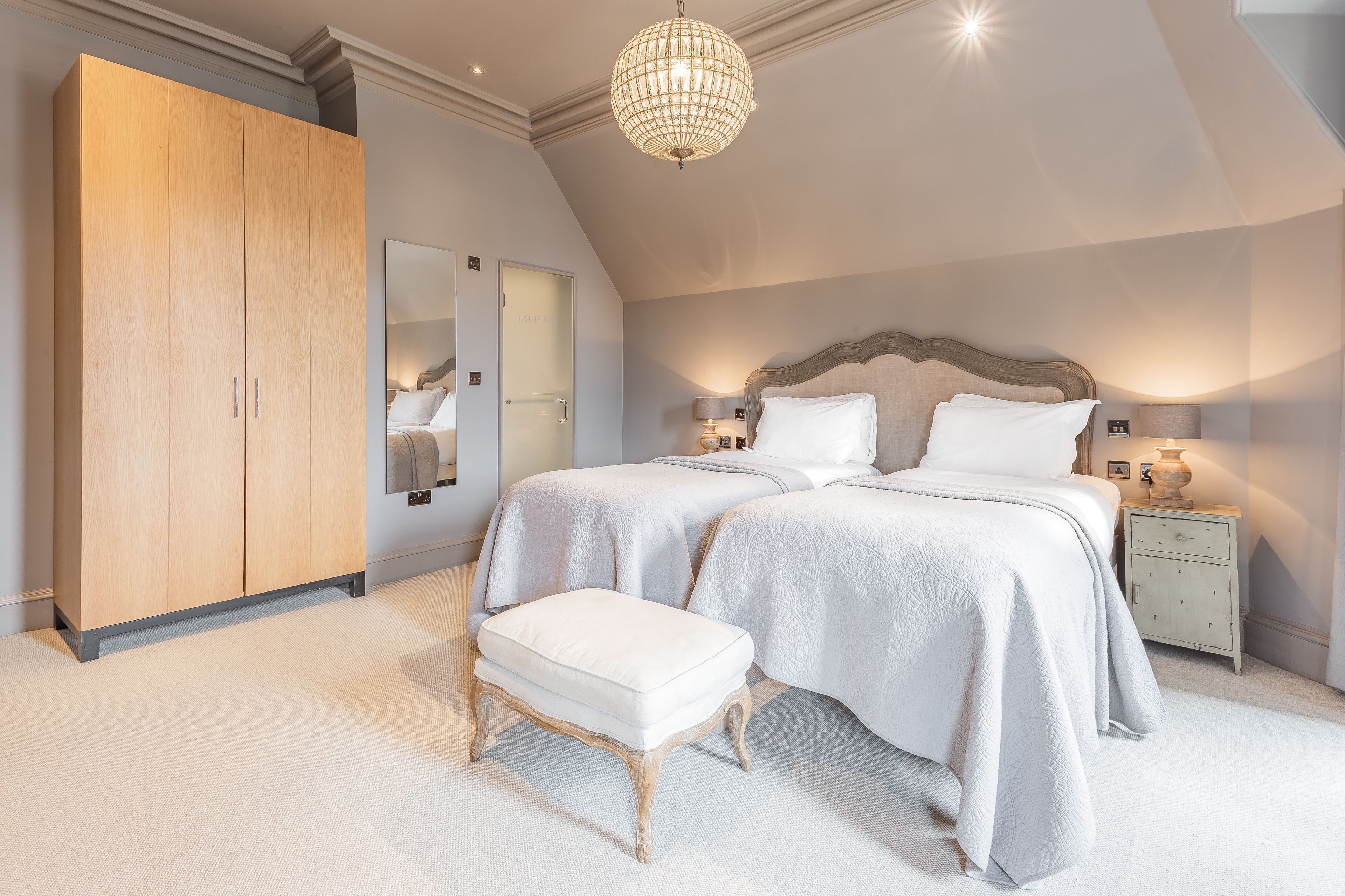 Une chambre avec deux lits, une armoire en bois, un banc, une table de chevet et un lustre.