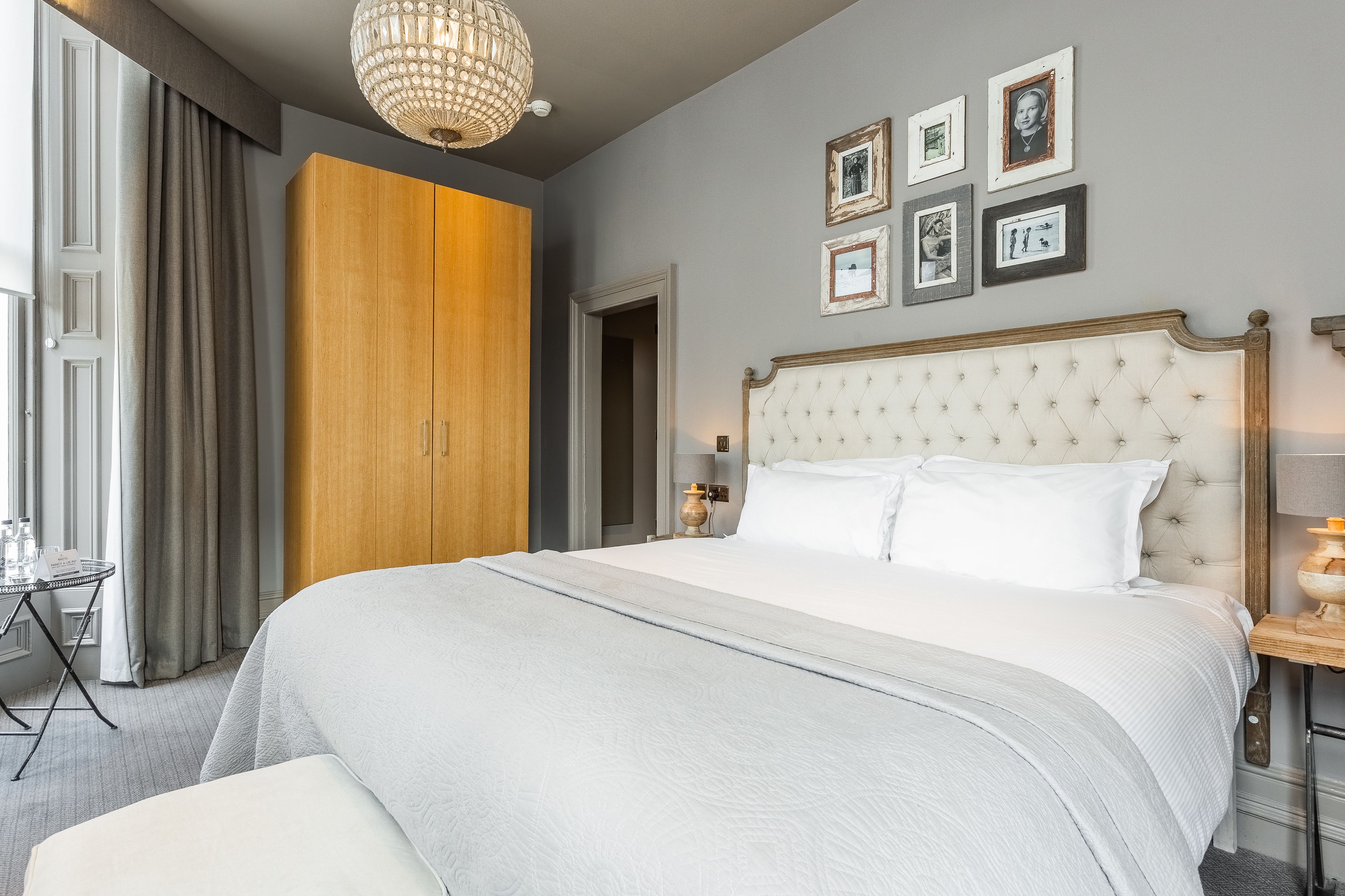 A neatly arranged hotel room with a large bed, a wooden wardrobe, a chandelier, and framed pictures on the wall.