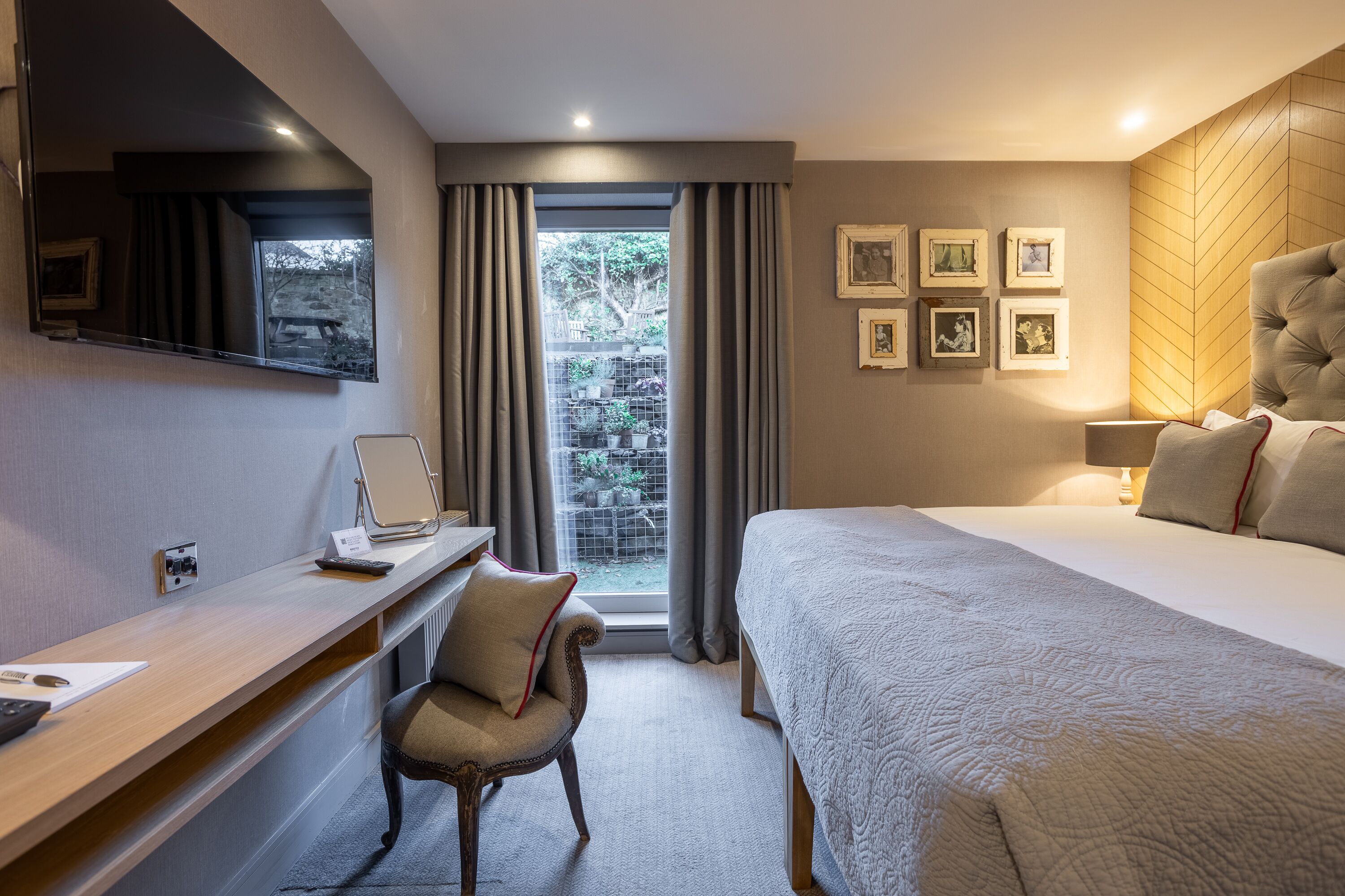 A hotel room with a large bed, a desk with a chair, a window with curtains, and framed pictures on the wall.
