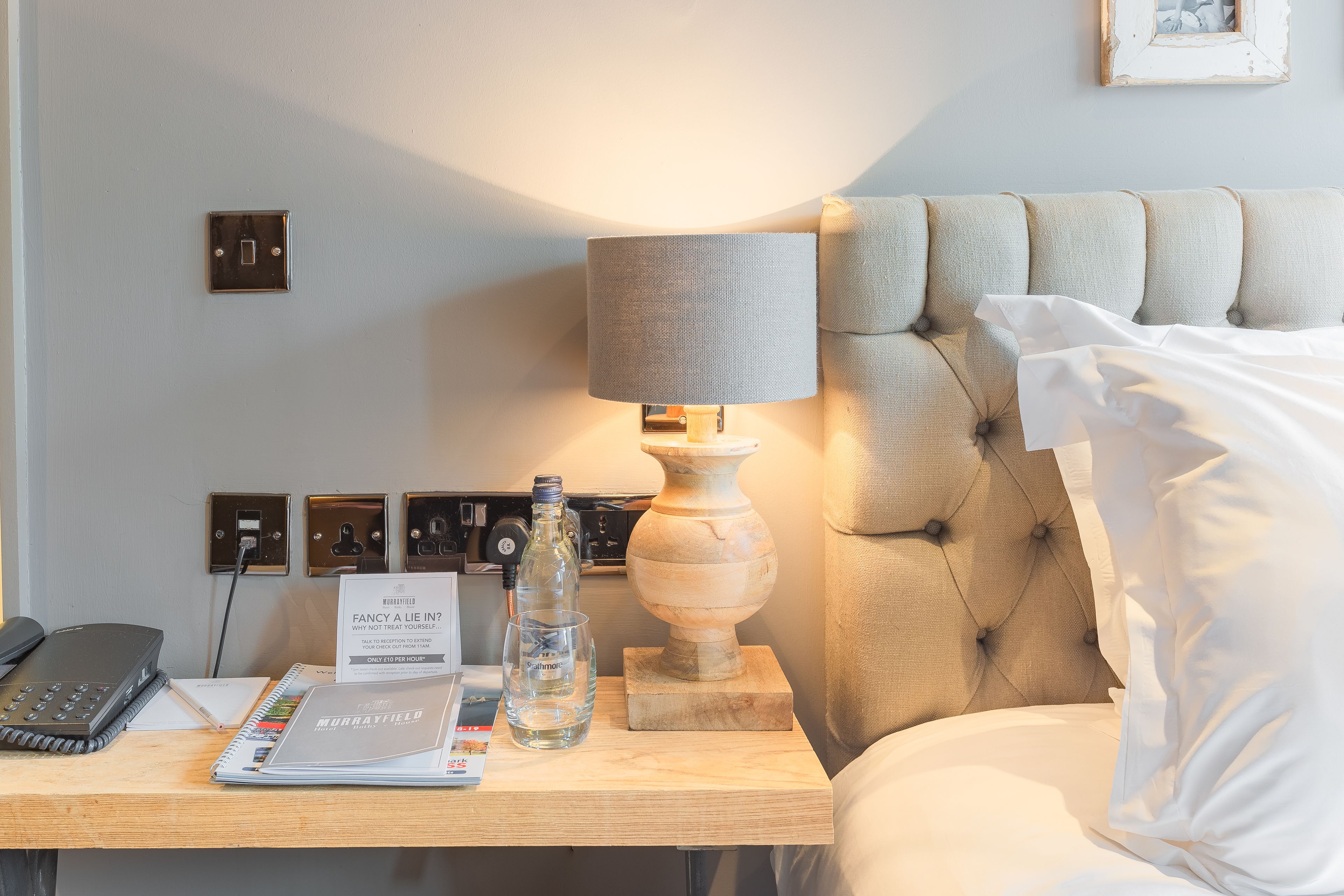 Et hotelværelse med en seng, et natbord med lampe, telefon, notesbog og en brochure.