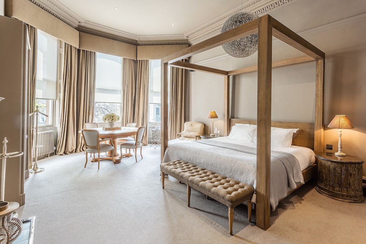 Ein Himmelbett mit Sitzbank, ein Esstisch mit Stühlen und eine Lampe auf einem Beistelltisch in einem hellen Raum mit großen Fenstern.