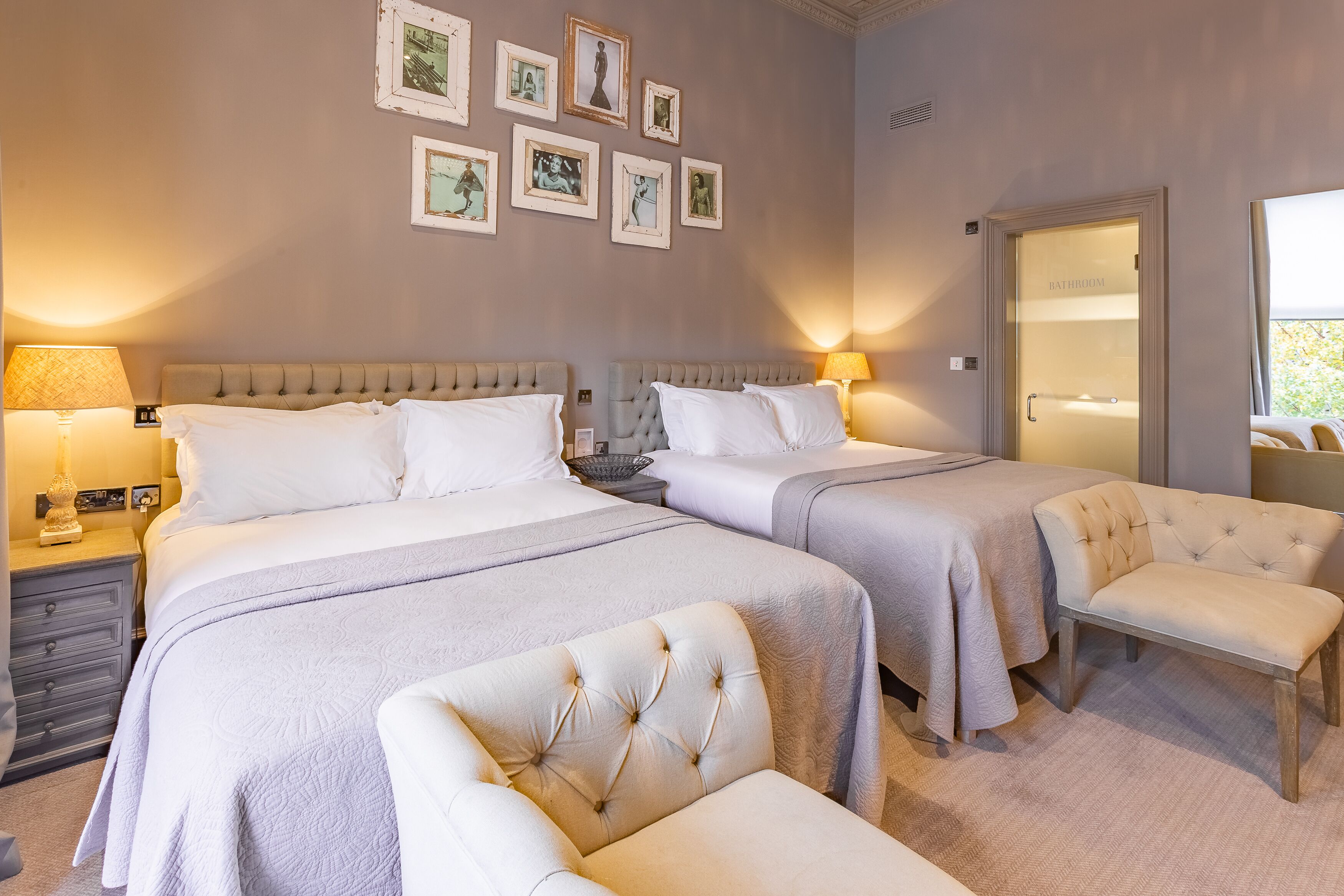 Ein Hotelzimmer mit zwei Betten, einer Bank, einem Nachttisch, einer Lampe und gerahmten Bildern an der Wand.