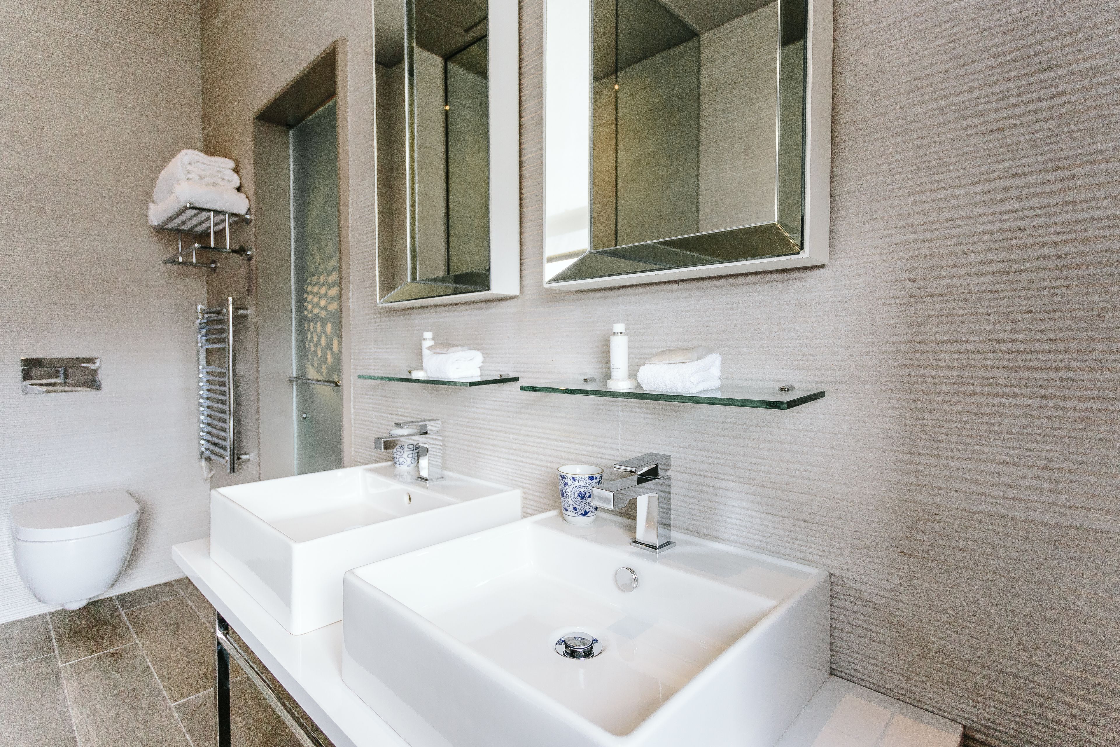 A modern bathroom with two sinks, a toilet, and a glass shelf with toiletries.