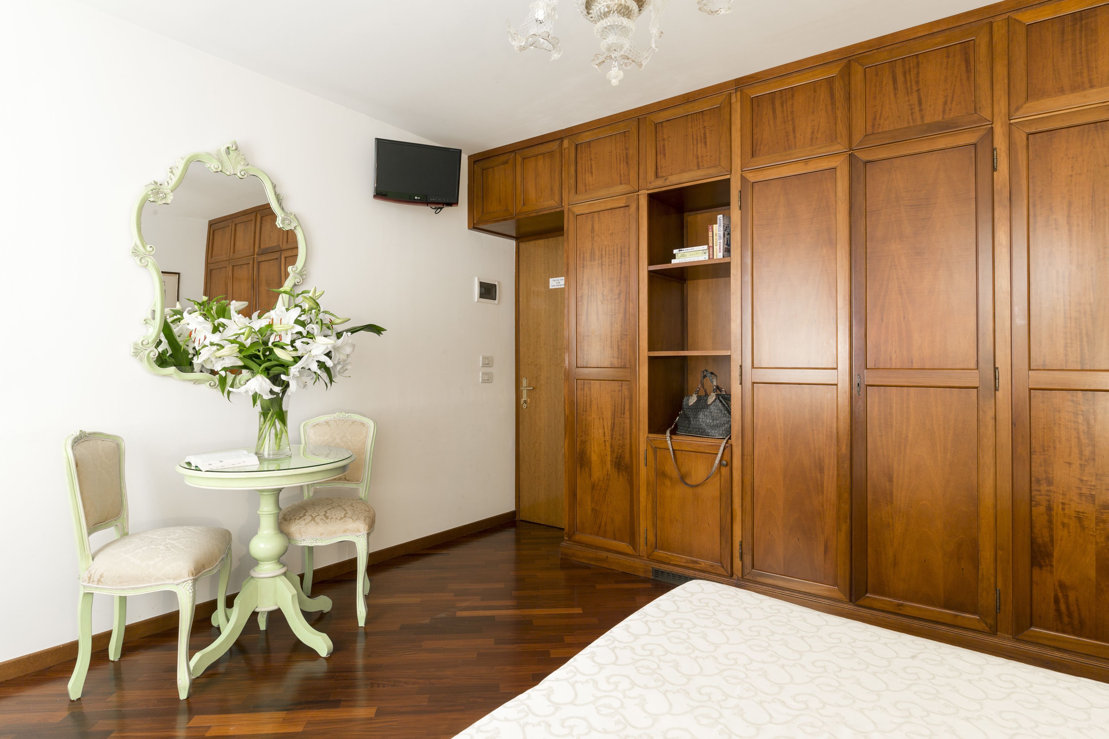 A room with a wooden wardrobe, a round table with chairs, a mirror, and a vase of flowers.