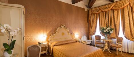 Habitación de hotel con una cama grande, dos mesitas de noche, una silla, una pequeña mesa con un jarrón de flores y un techo de madera con vigas a la vista.