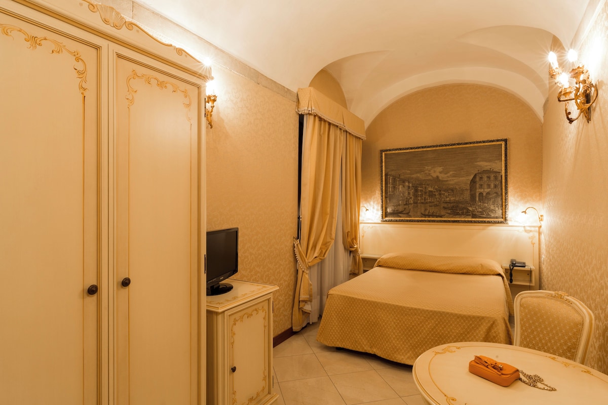 Quarto de hotel com cama, televisão sobre um móvel, escrivaninha com telefone e quadro na parede.