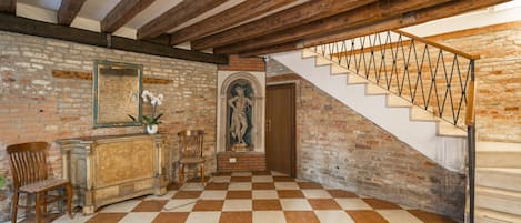 Una habitación con piso a cuadros, vigas de madera, una escalera con barandilla de metal, una estatua y una pared de ladrillo.