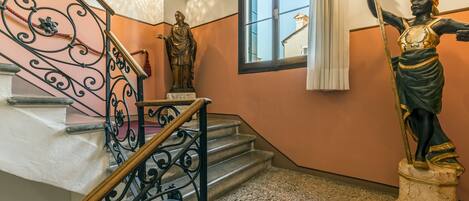 A staircase with ornate metal railings, a statue of a figure holding a staff, and a window with a view of buildings outside.