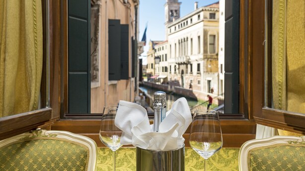 A table with two wine glasses and a bottle of champagne, set against a window overlooking a canal with historic buildings. - Palazzo Guardi (Venice)