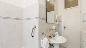 Un baño con un lavabo blanco, un estante de vidrio con artículos de aseo, un perchero y una zona de ducha.