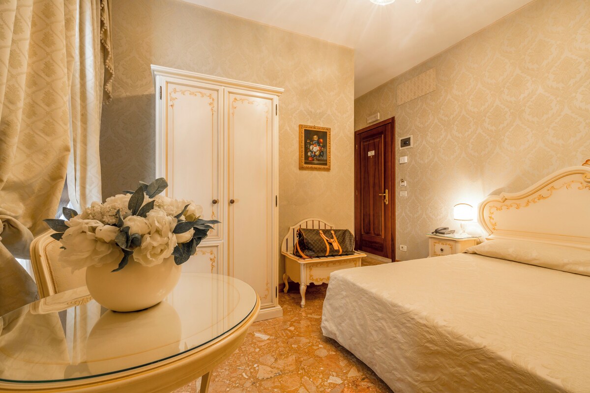 Habitación de hotel con una cama, una mesita de noche con lámpara, una silla, un armario blanco y un jarrón con flores.