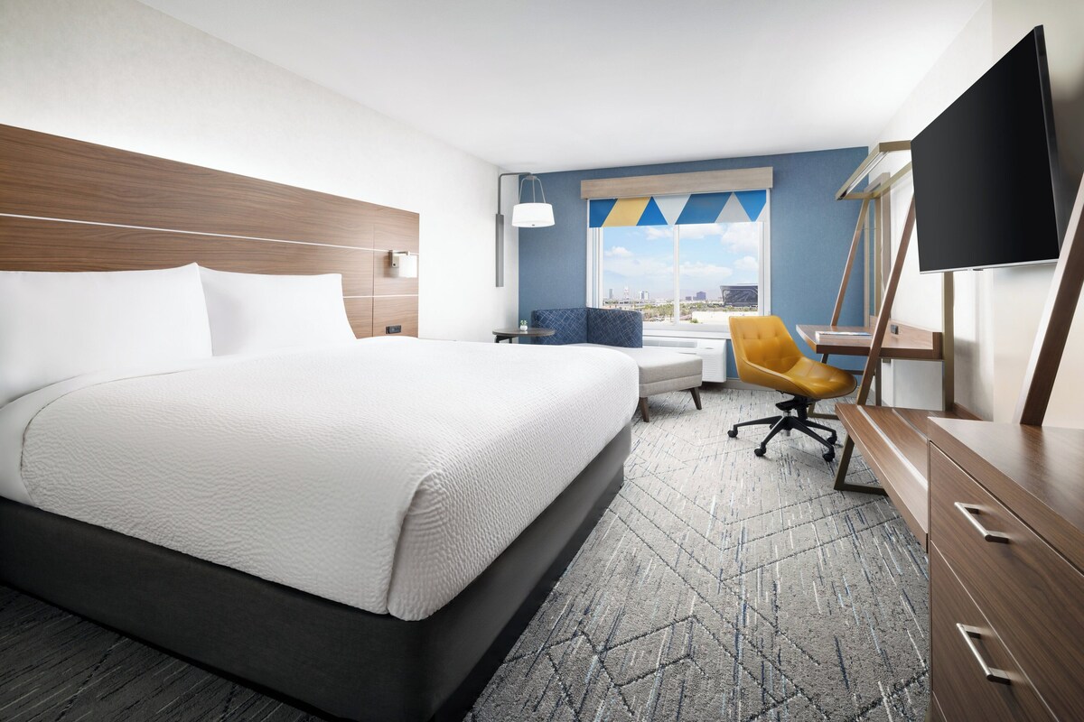Habitación de hotel con una cama grande, un escritorio, una silla, un televisor y una ventana con vista a la ciudad.