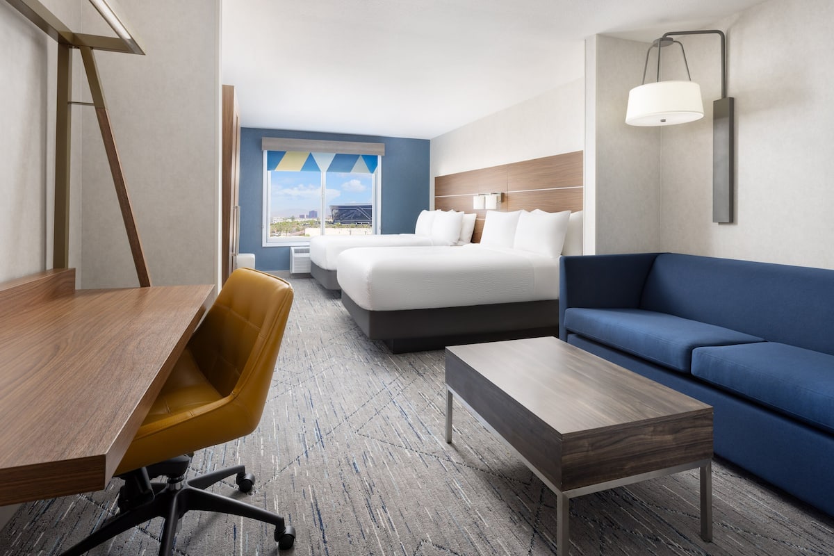 A hotel room with a wooden desk, a blue sofa, a yellow chair, a bed with white bedding, and a view of the city through a window.